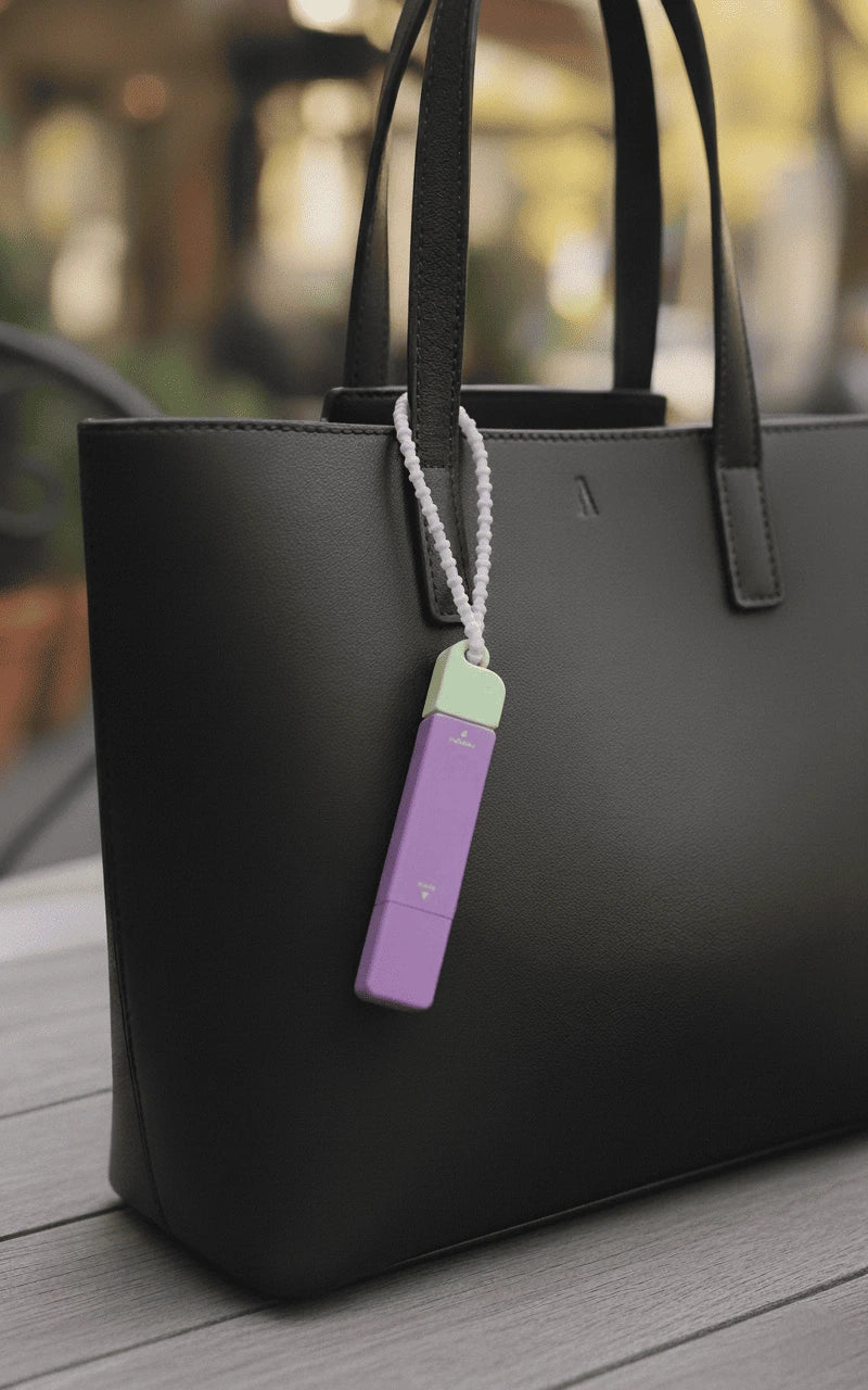 A black handbag sits on a wooden table, adorned with a purple and green portable stain remover attached to its handle. The scene is bright, with a blurred outdoor background.