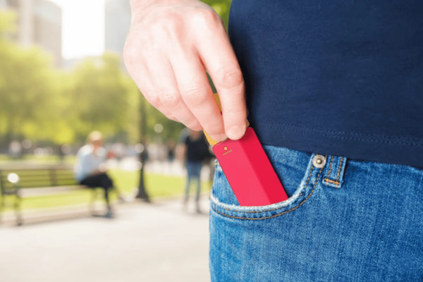 A person in blue jeans and a navy shirt slips a red portable stain remover into their pocket. The background shows a sunny park with trees and blurred people.