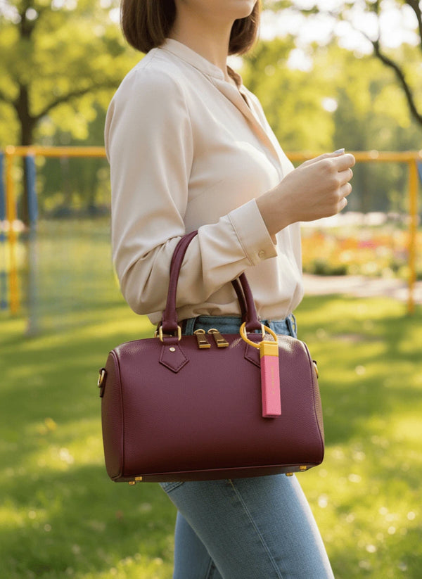 A woman in a beige blouse and jeans holds a burgundy handbag with a pink portable stain remover, standing in a sunlit park with green grass and blurry trees.